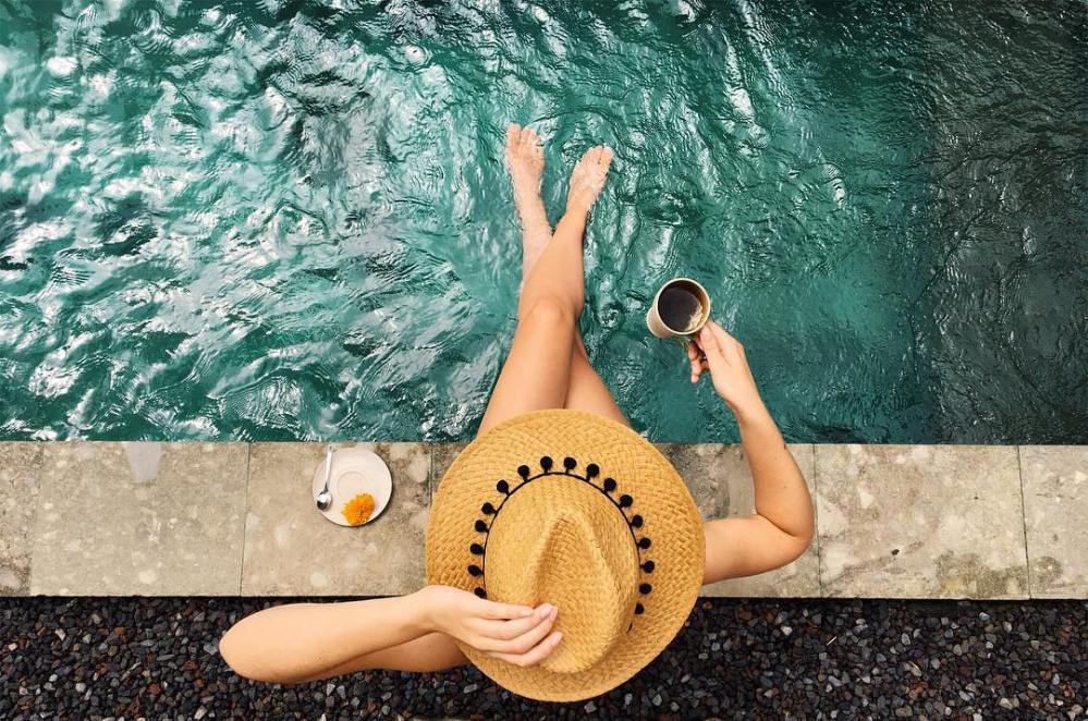 Woman sits with her coffee and hat on the edge of a pool in Ubud, Bali